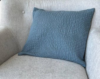 May include: A square, textured, blue decorative pillow rests on a light gray armchair. The pillow has a quilted pattern and a soft, inviting appearance. The chair has a button detail on the backrest, and the background includes greenery and a decorative lantern.