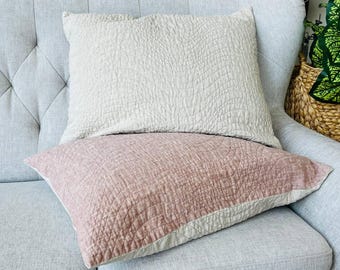 May include: Two decorative throw pillows on a light gray upholstered chair. The top pillow is a light beige color, and the bottom pillow is a dusty rose color. Both pillows have a textured, quilted pattern.
