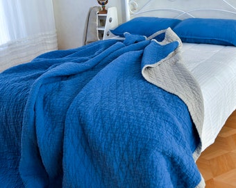 May include: A blue and gray reversible quilt with a textured pattern. The quilt is folded on a white bed with blue and gray pillows.