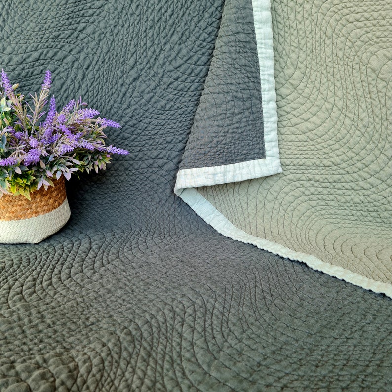 May include: A gray and beige quilted throw blanket with a wavy pattern. The blanket is folded in half and has a white border.