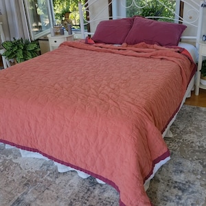 May include: A coral-colored quilted bedspread with a white ruffle and a burgundy trim. The bedspread is on a white bed with two burgundy pillows.