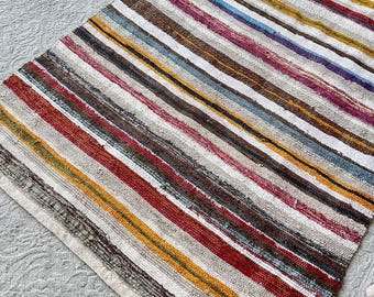May include: A rectangular, handwoven rug with a striped pattern. The rug features a variety of colors, including red, brown, yellow, blue, and white. The stripes run lengthwise across the rug, creating a visually appealing design. The rug is placed on a light gray surface.