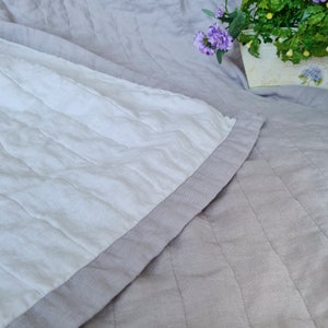 Puede incluir: Primer plano de una colcha acolchada con un centro blanco y un borde gris. La colcha est&aacute; hecha de una tela suave y texturizada. El fondo es gris claro, con un toque de arreglo floral.
