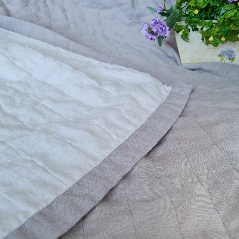 Puede incluir: Primer plano de una colcha acolchada con un centro blanco y un borde gris. La colcha est&aacute; hecha de una tela suave y texturizada. El fondo es gris claro, con un toque de arreglo floral.