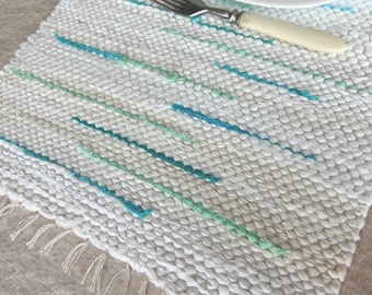 May include: A rectangular woven placemat in off-white featuring horizontal dashed lines in shades of teal and mint green. The placemat has a textured surface with fringe along the bottom edge. A white plate, a fork, and a folded napkin are placed on top.
