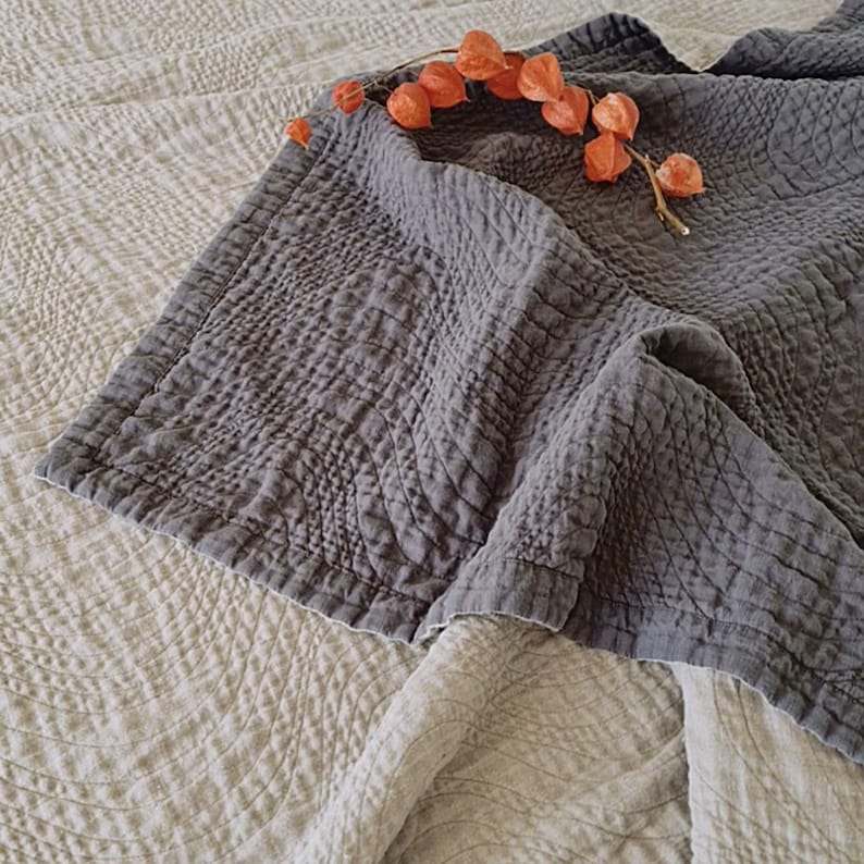 May include: A gray quilted throw blanket with a textured pattern. The blanket is folded and draped over a white textured blanket.