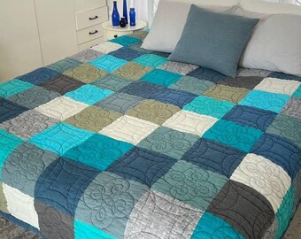 May include: A quilted bedspread with a patchwork design in shades of blue, teal, gray, and white. The squares are quilted with a floral pattern. The bed is made up with pillows and a white metal bed frame.