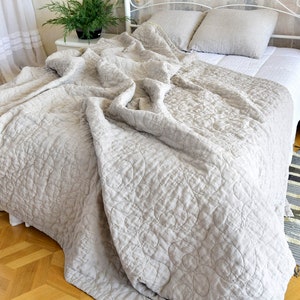 Puede incluir: Una colcha de lino gris claro con un patrón acolchado de círculos. La colcha está drapeada sobre una cama con sábanas y almohadas de lino blanco.