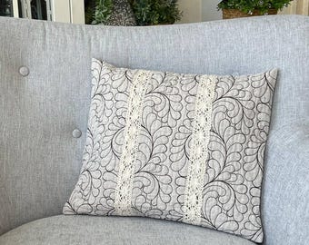 May include: A square throw pillow featuring a light beige fabric base with a repeating black stitched feather pattern. The pillow is accented by two vertical strips of cream-colored lace trim running down the center, creating a textured design.