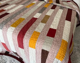 May include: A handmade quilt with a patchwork design in shades of pink, red, yellow, brown, and white. The quilt is draped over a bed with matching pillows. The quilt's dimensions are not visible in the image.