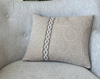 May include: A square decorative pillow with a neutral beige tone, featuring a central vertical band of white and brown lace. The pillow has a quilted texture with a subtle floral pattern, adding a touch of elegance to home decor.