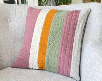 May include: A square decorative pillow with vertical stripes in shades of pink, white, orange, and green. The pillow has a quilted design with curved stitching. The pillow is resting on a gray upholstered armchair.