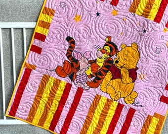 May include: Pink baby quilt featuring Winnie the Pooh, Tigger, and Piglet. The quilt has a pink background with embroidered stars and swirls. Vertical stripes in red, yellow, and orange border the quilt. The quilt is placed on a crib.