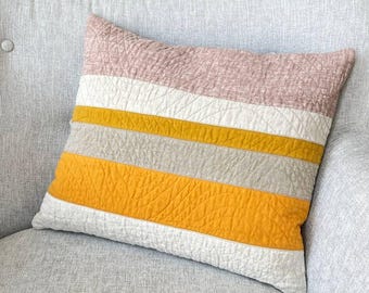May include: A square decorative pillow with horizontal stripes in shades of pink, white, gold, and gray. The pillow has a quilted texture and is resting on a gray upholstered sofa. Home decor.