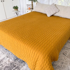 K&ouml;nnte beinhalten: Eine senfgelbe gesteppte Tagesdecke mit einem wellenf&ouml;rmigen Muster. Die Decke liegt auf einem Bett mit wei&szlig;en Kissen und einem wei&szlig;en Kopfteil.