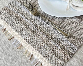 May include: A woven placemat with a textured, gray and beige pattern. A white border with fringe accents the edge. A white plate, a folded napkin, and a fork are on the placemat. The napkin is tied with a string.