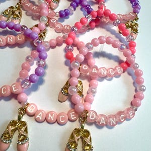 May include: A collection of pink, purple, and clear beaded bracelets. Some bracelets spell out "DANCE" with white letter beads. Each bracelet has a charm of a pink ballet slipper with gold accents and rhinestones.