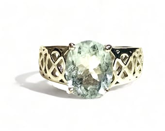 May include: A gold ring with a delicate, intricate design featuring a large oval-shaped light green gemstone.