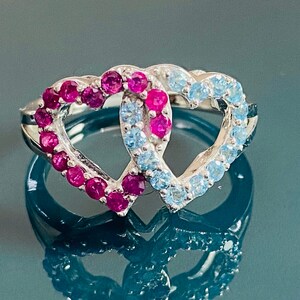Puede incluir: Anillo de plata con dos formas de corazón entrelazadas. Un corazón está adornado con pequeñas piedras rosas vibrantes, el otro con piedras azul claro. El anillo se muestra sobre una superficie reflectante.