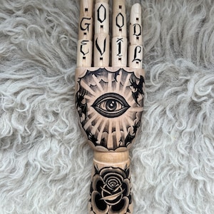 May include: A wooden hand with articulated fingers painted with a black and white tattoo design. The tattoo features an all-seeing eye with rays of light, clouds, and birds. The fingers spell out "GOOD EVIL". The bottom of the hand is painted with a black rose.