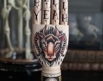 Hölzerne Hand mit Ziegen- und Totenkopf-Tattoo-Design