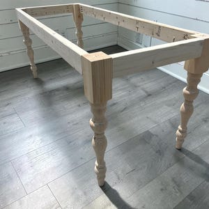 May include: A rectangular unfinished wooden table frame featuring four decorative turned legs with a bulbous design. The structure is constructed from light-colored pine boards joined at the corners with visible pocket hole screws along the inner rails.