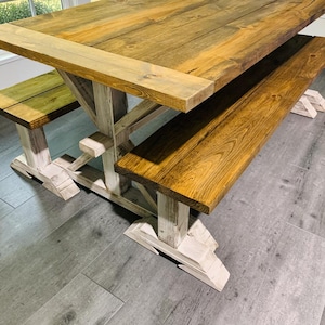 Könnte beinhalten: Ein rechteckiger Esstisch mit einer naturfarbenen braunen Holzmaserung und einem weiß gewischten Gestell, begleitet von zwei passenden Bänken mit braunen Holzsitzen und weiß gewischten Beinen, die eine stabile Kreuzstreben-Stützstruktur aufweisen.