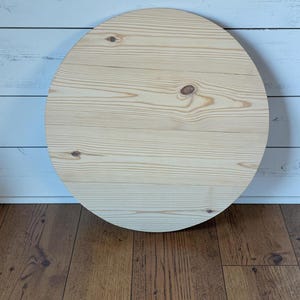 May include: A circular wooden board made of light-colored pine planks. The surface features a natural wood grain pattern with visible knots and horizontal lines where the individual boards are joined together to form a smooth, round shape.