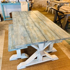 May include: A rustic farmhouse-style dining table and bench set. The table features a light blue-gray stained wooden top with visible wood grain. The base and bench legs are painted white. The set is displayed in a showroom setting.