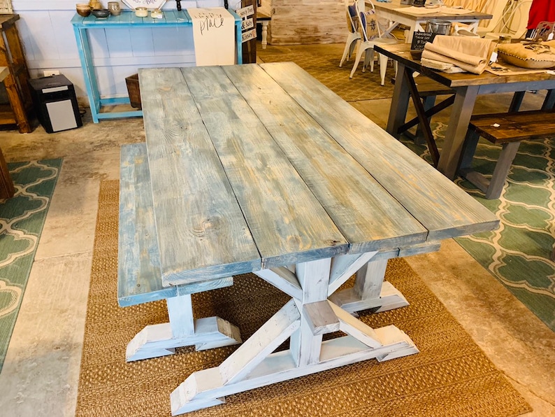May include: A rustic farmhouse-style dining table and bench set. The table features a light blue-gray stained wooden top with visible wood grain. The base and bench legs are painted white. The set is displayed in a showroom setting.