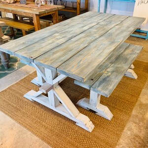 May include: A large, rustic farmhouse-style dining table with a matching bench. The tabletop is made of weathered wood with a blue-gray finish, supported by white, X-shaped legs. The table sits on a woven jute rug.