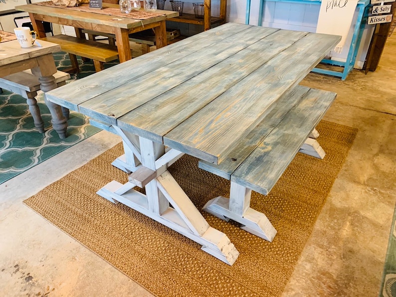 May include: A large, rustic farmhouse-style dining table with a matching bench. The tabletop is made of weathered wood with a blue-gray finish, supported by white, X-shaped legs. The table sits on a woven jute rug.
