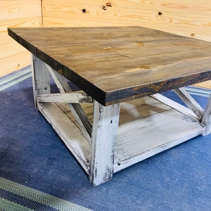 May include: A square coffee table featuring a dark brown stained wooden top and a distressed white painted base. The base includes four sturdy legs connected by X-shaped cross-bracing on the sides and a lower shelf for additional storage space.