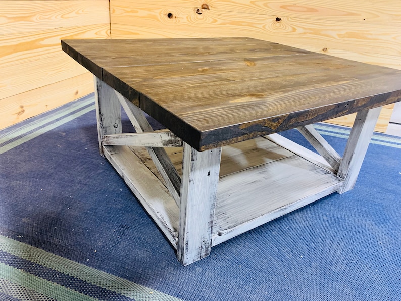 May include: A square coffee table featuring a dark brown stained wooden top and a distressed white painted base. The base includes four sturdy legs connected by X-shaped cross-bracing on the sides and a lower shelf for additional storage space.