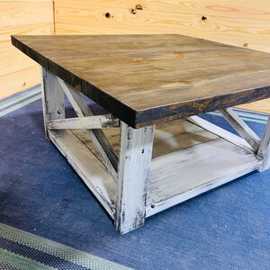 May include: A square coffee table featuring a dark brown stained wooden top and a distressed white painted base. The base includes four sturdy legs connected by X-shaped cross-bracing on the sides and a lower shelf for additional storage space.
