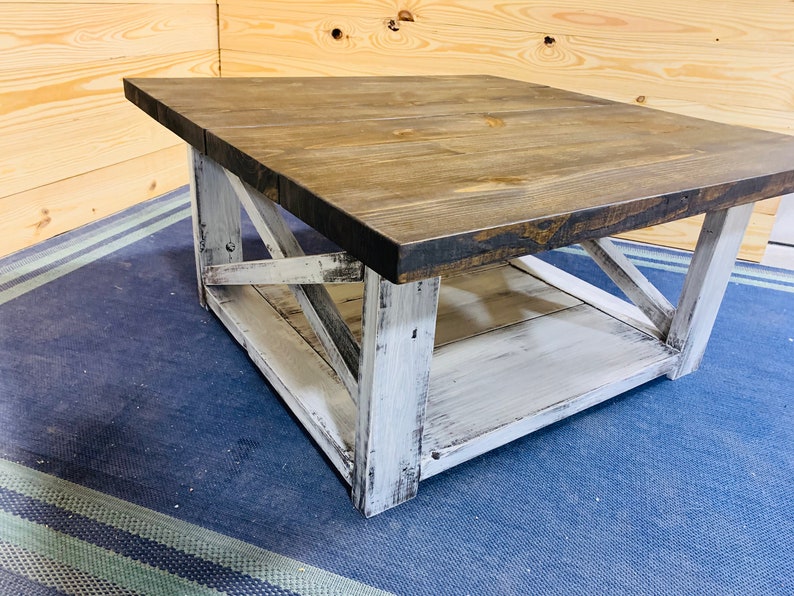 May include: A square coffee table featuring a dark brown stained wooden top and a distressed white painted base. The base includes four sturdy legs connected by X-shaped cross-bracing on the sides and a lower shelf for additional storage space.