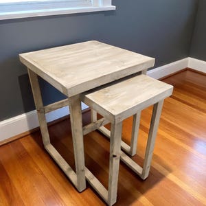 May include: Two light-colored wooden nesting tables. The larger table is square, with a smaller square table nested underneath. The tables have a rustic, weathered appearance, with visible wood grain. The tables are placed on a hardwood floor.