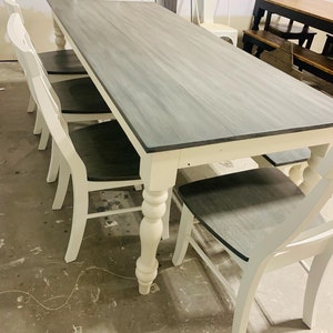 K&ouml;nnte beinhalten: Ein rechteckiger Esstisch mit einer grau gebeizten Holzplatte und wei&szlig; lackierten Beinen im gedrechselten Design. Der Tisch wird von mehreren passenden St&uuml;hlen mit wei&szlig;em Rahmen und dunkelgrauen Holzsitzfl&auml;chen erg&auml;nzt.