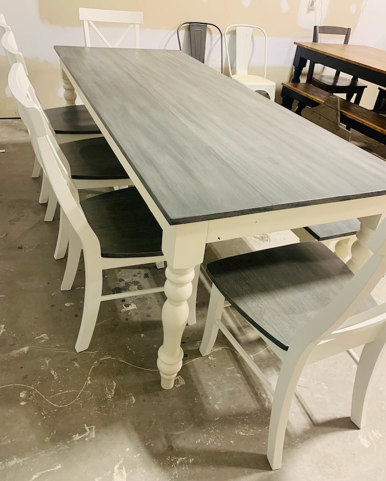 K&ouml;nnte beinhalten: Ein rechteckiger Esstisch mit einer grau gebeizten Holzplatte und wei&szlig; lackierten Beinen im gedrechselten Design. Der Tisch wird von mehreren passenden St&uuml;hlen mit wei&szlig;em Rahmen und dunkelgrauen Holzsitzfl&auml;chen erg&auml;nzt.