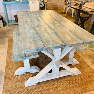 May include: A large, rustic wooden dining table with a matching bench. The tabletop has a weathered blue-gray finish, while the base and bench legs are painted white. The table is supported by a sturdy, X-shaped base.