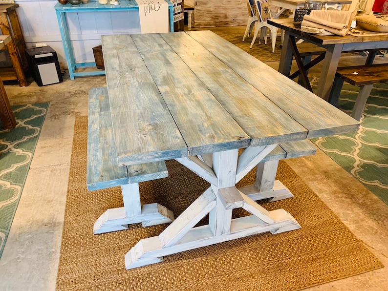 May include: A large, rustic wooden dining table with a matching bench. The tabletop has a weathered blue-gray finish, while the base and bench legs are painted white. The table is supported by a sturdy, X-shaped base.