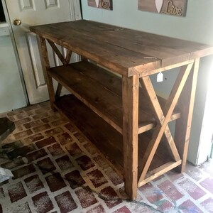 May include: A rectangular dark brown wooden console table featuring a flat top surface, one lower shelf, and X-shaped support braces on both side panels. The furniture piece is constructed from thick wooden planks with a visible grain texture.