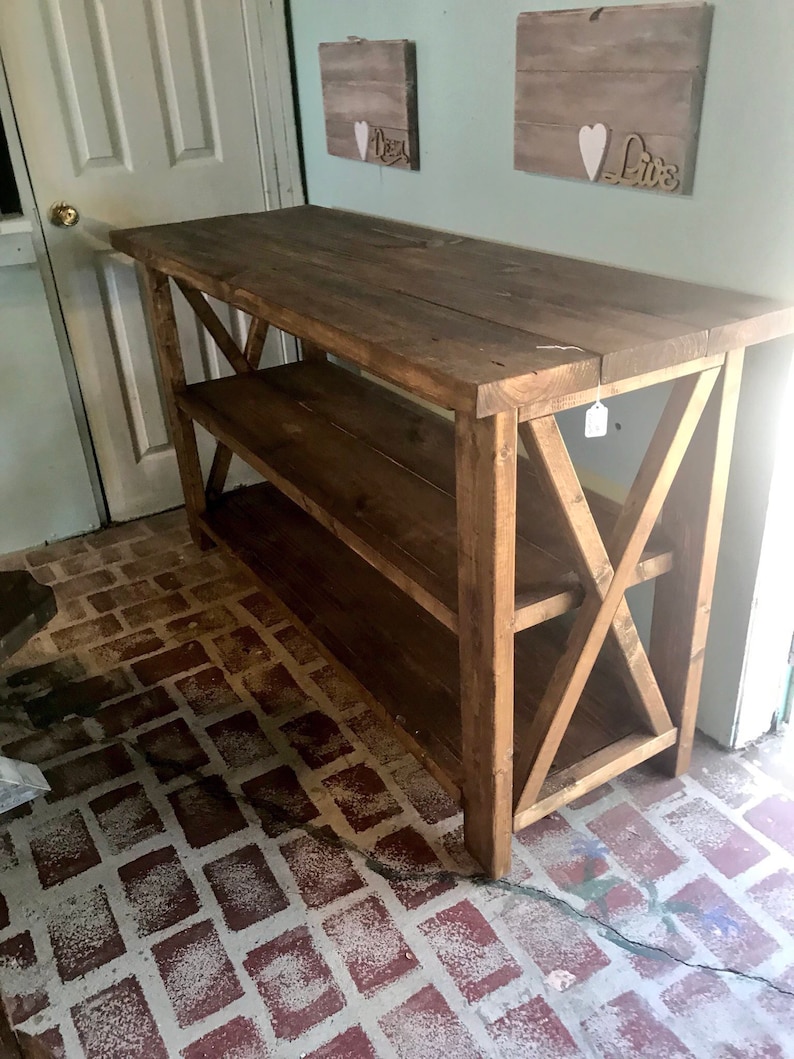 May include: A rectangular dark brown wooden console table featuring a flat top surface, one lower shelf, and X-shaped support braces on both side panels. The furniture piece is constructed from thick wooden planks with a visible grain texture.
