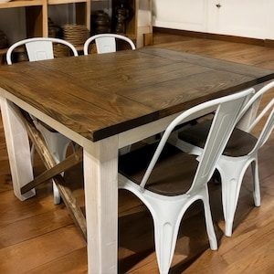 May include: A rustic dining table with a dark wood top and white painted legs. The table is surrounded by three white metal chairs with dark wood seats. The table is approximately 48 inches square.