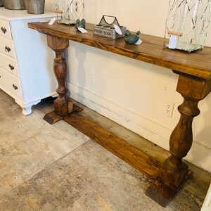 May include: A long, rectangular console table made of dark brown stained wood. The table features a flat top surface supported by two thick, turned pedestal legs connected by a horizontal base board that runs along the floor between them.