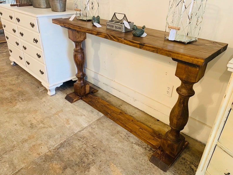 May include: A long, rectangular console table made of dark brown stained wood. The table features a flat top surface supported by two thick, turned pedestal legs connected by a horizontal base board that runs along the floor between them.