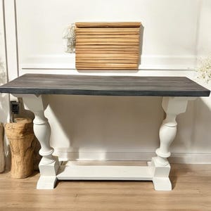 May include: A rectangular console table featuring a dark gray, weathered-look wooden top supported by two white, turned pedestal-style legs connected by a horizontal white base stretcher. The table has a clean, contrasting two-tone finish.