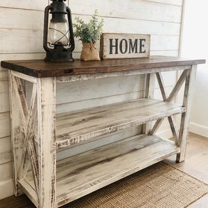May include: A wooden console table with a dark brown top and a distressed white base featuring X-shaped side supports and two open shelves. On the surface sits a black metal lantern, a small potted green plant, and a rectangular sign reading HOME.