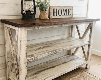 May include: A wooden console table with a dark brown top and a distressed white base featuring X-shaped side supports and two open shelves. On the surface sits a black metal lantern, a small potted green plant, and a rectangular sign reading HOME.