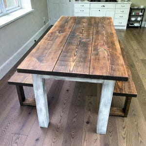 May include: A rustic wooden dining table and bench set. The tabletop is made of wide, stained planks with visible wood grain. The legs are painted white, contrasting with the dark brown tabletop. The bench mirrors the table's design, creating a cohesive look.